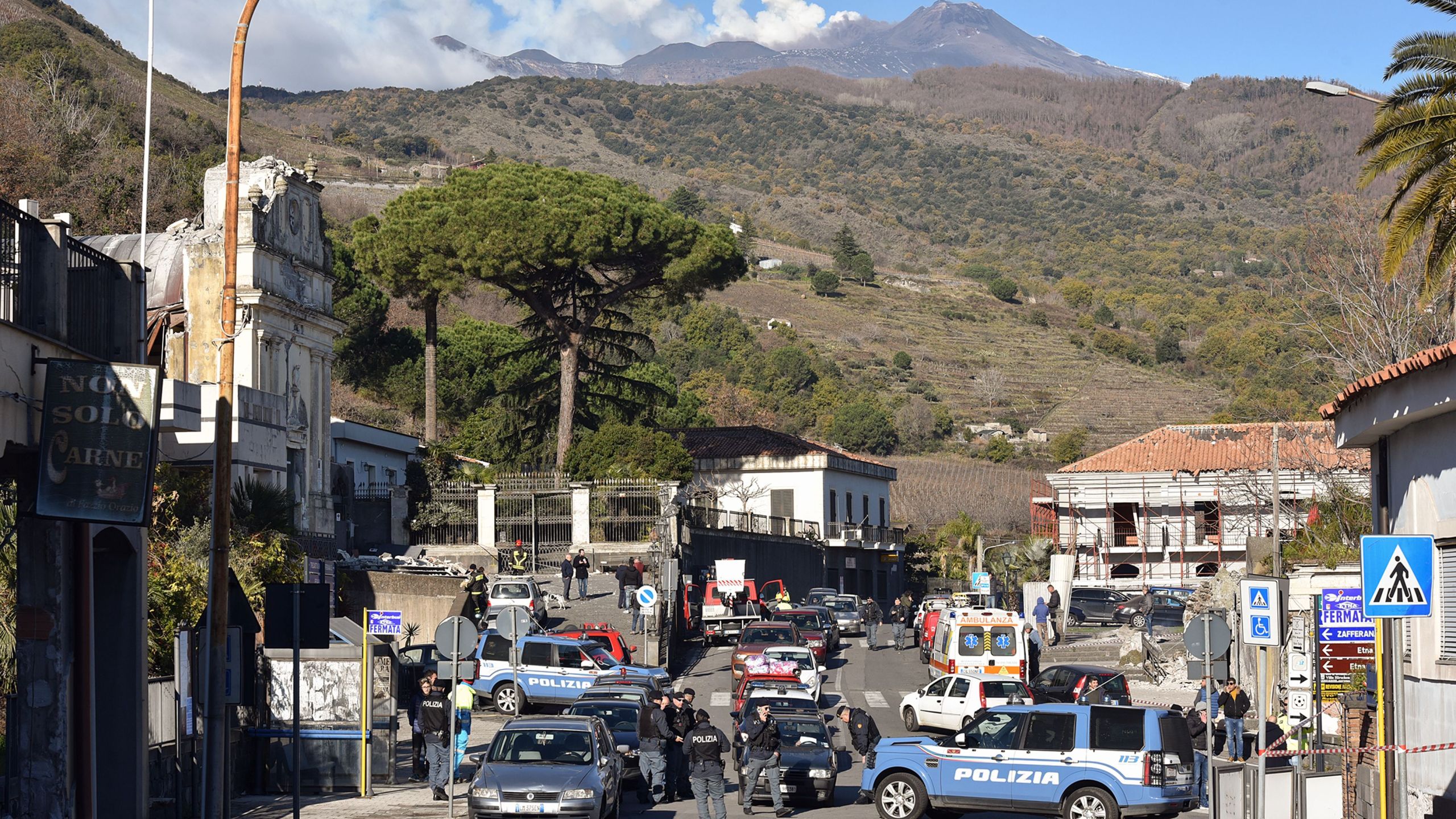 A general view shows police officers and an ambulance outside the damaged church of Parrocchia Maria Ss.Del Rosario (left), with smoke rising from Mount Etana in Zafferana Etnea near Catania on Dec. 26, 2018. (Credit: Giovanni Isolino/AFP/Getty Images)