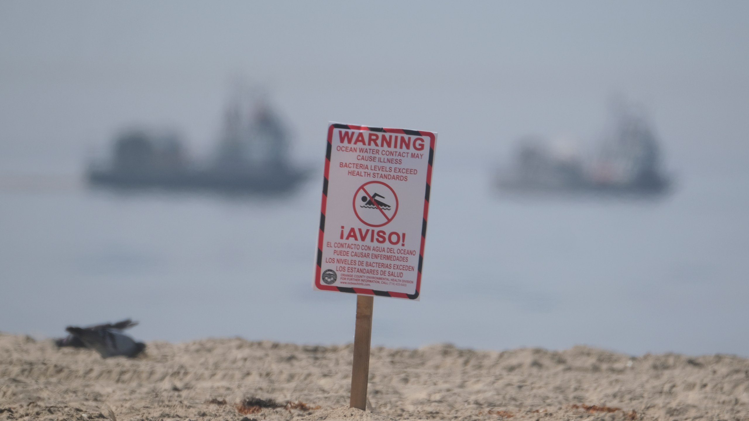 A posted sign warning about water contact may cause illness is posted by life guards after an oil spill in Huntington Beach, Calif., Sunday, Oct. 3, 2021. Vessels with the Marine Spill Response Corporation (MSRC) vessel, background, an oil spill removal organization (OSRO), deploy floating barriers around the largest oil spills in recent Southern California history fouled popular beaches and killed wildlife while crews scrambled Sunday to contain the crude before it spread further into protected wetlands. (AP Photo/Ringo H.W. Chiu)