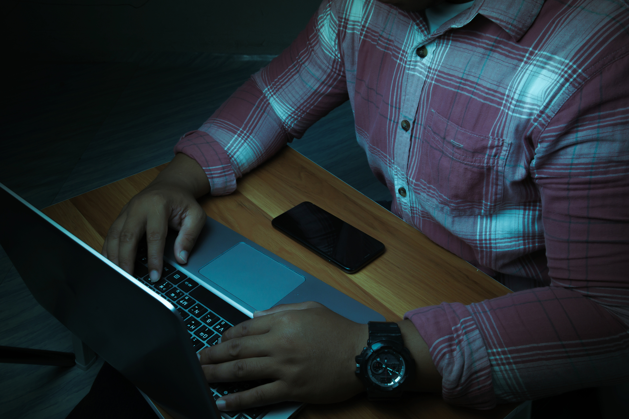 A man is sitting on a laptop in a dark room with lights shining down on it.