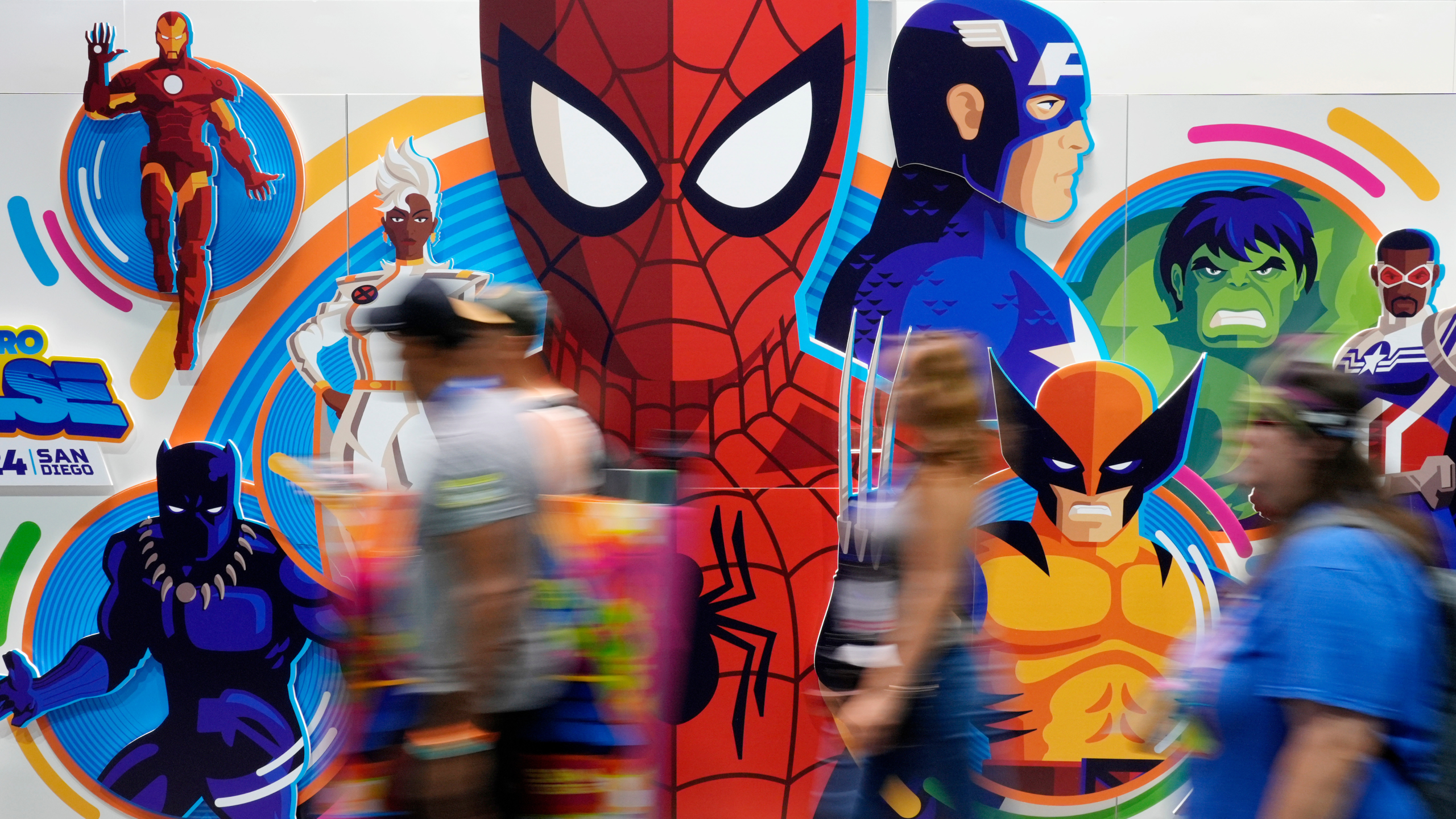 Convention attendees blur past a wall of illustrated Marvel superhero characters.