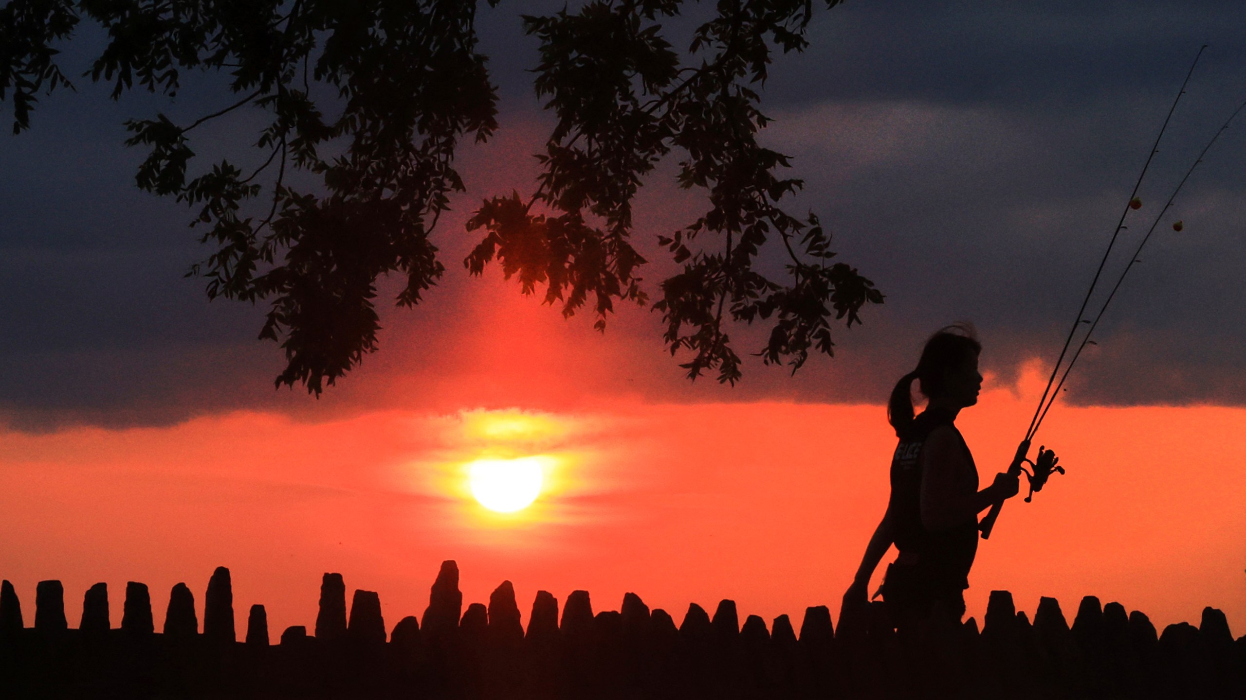 A person walks with fishing gear as the sun is obscured from the wildfires in the western United States and Canada in Oconomowoc, Wis. Saturday, Aug. 3, 2024. (John Hart/Wisconsin State Journal via AP)