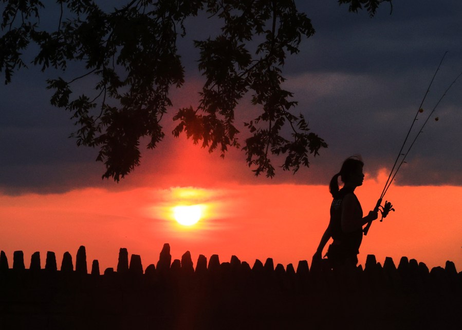 A person walks with fishing gear as the sun is obscured from the wildfires in the western United States and Canada in Oconomowoc, Wis. Saturday, Aug. 3, 2024. (John Hart/Wisconsin State Journal via AP)