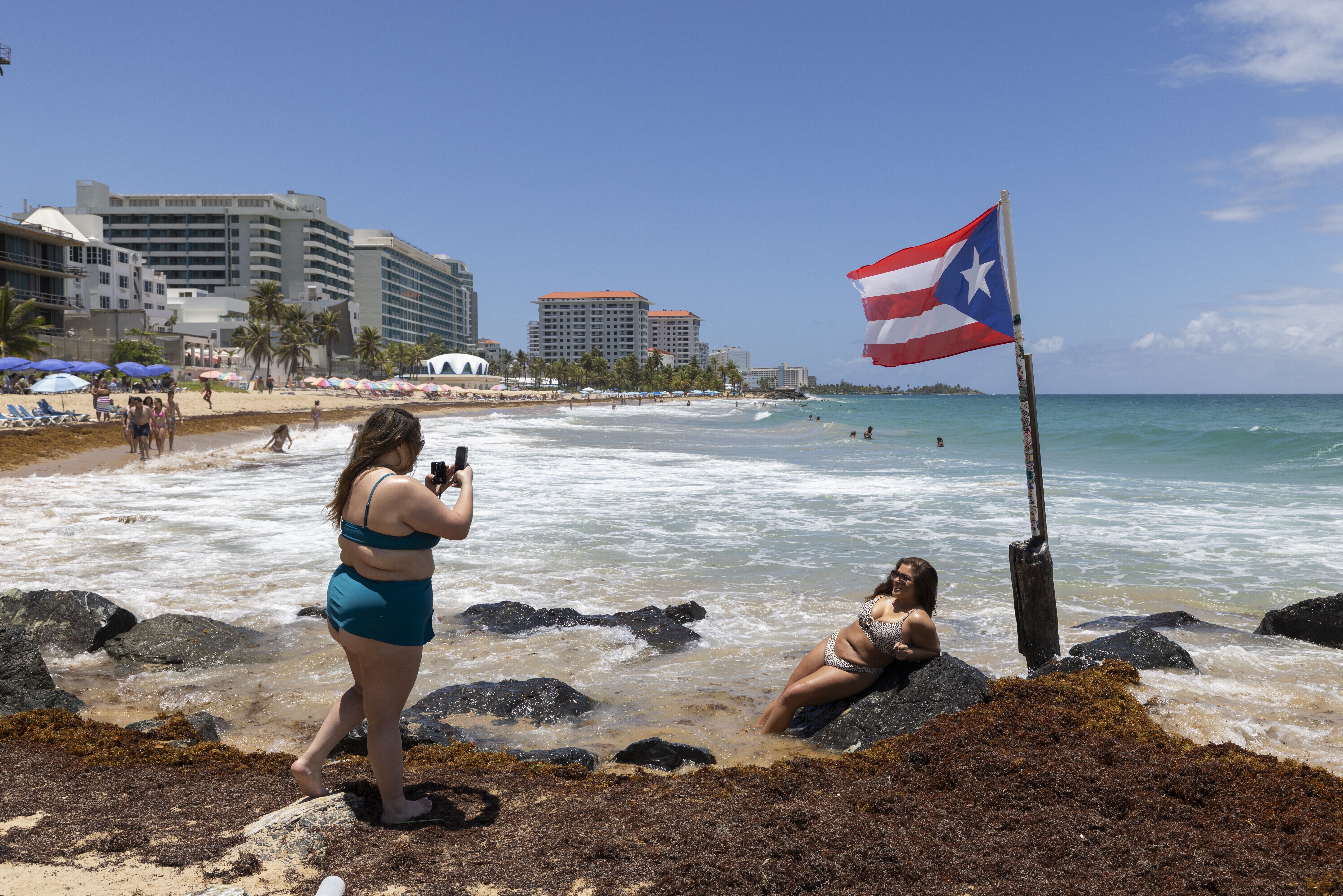A woman poses for photos next to a Puerto Rican flag along the beach in Condado, Puerto Rico, as Hurricane Erin approaches, Friday, Aug. 15, 2025. (AP Photo/Alejandro Granadillo)